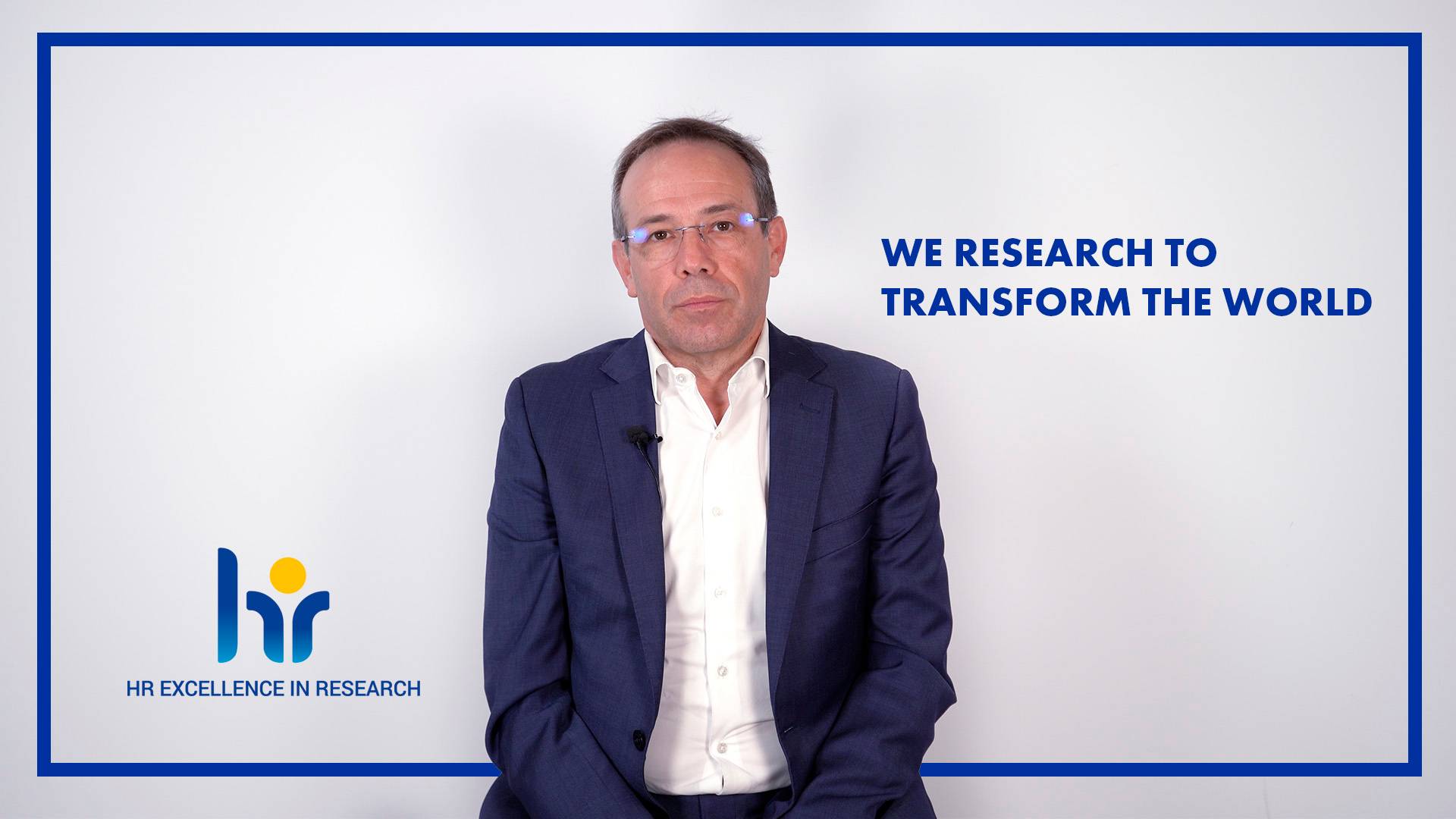Hombre en traje con fondo blanco y texto "We research to transform the world" en una campaña de la Universidad de Comillas sobre investigación y excelencia en investigación.