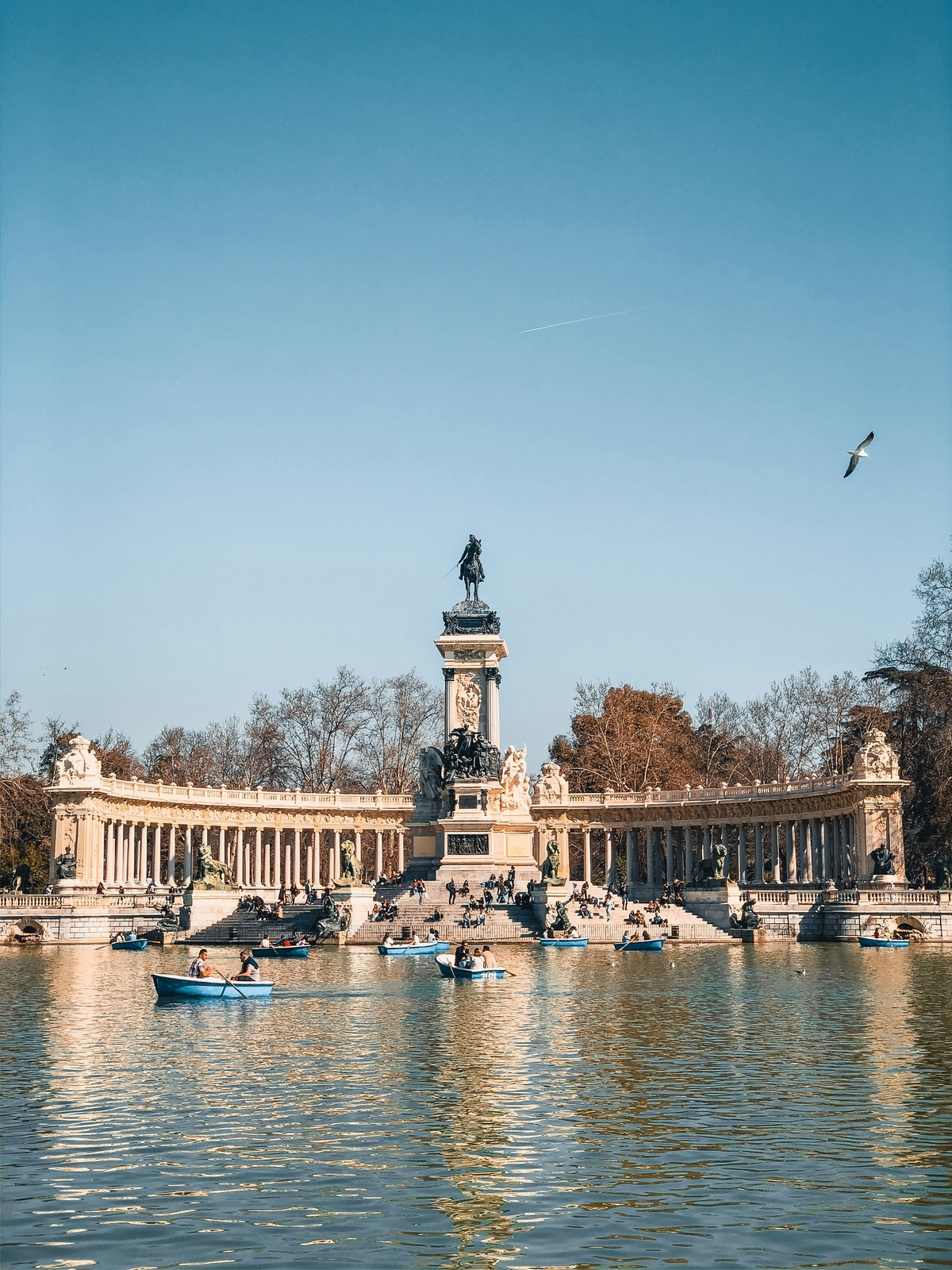 Una imagen del Palacio de Cristal en un parque, rodeado de árboles y con personas en botes sobre el agua.