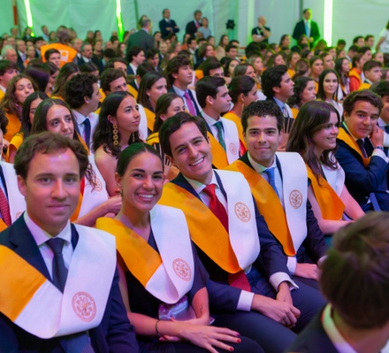 A group of young graduates wearing academic sashes and robes at a graduation ceremony, smiling and looking towards the stage.