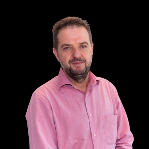 A professional portrait of a smiling man with a beard, wearing a pink shirt, against a black background.