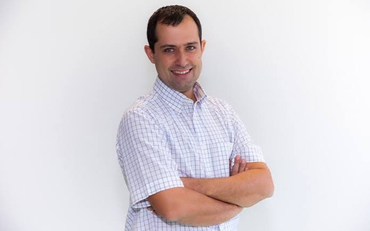 Un hombre sonriente, vestido con una camisa a cuadros, posa con los brazos cruzados en un fondo blanco.
