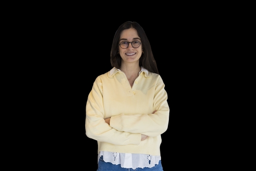 Una mujer sonriente con un suéter amarillo y gafas, de pie con los brazos cruzados sobre un fondo negro.