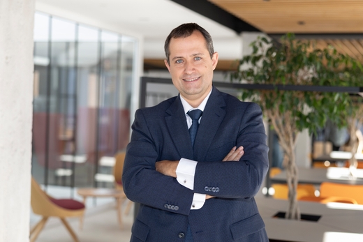 Un hombre sonriente está de pie en un entorno moderno y luminoso, con las manos cruzadas sobre el pecho.