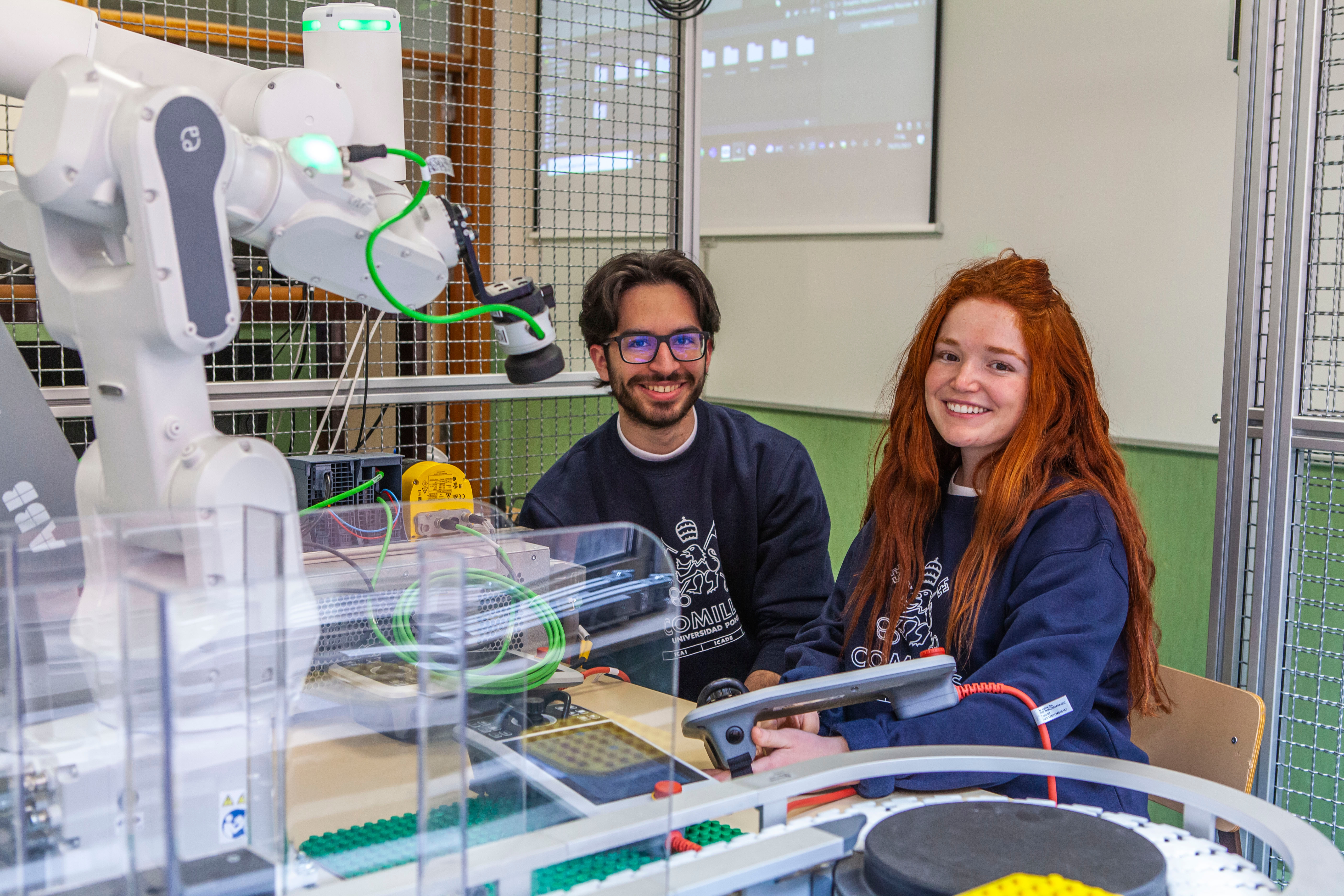 Dos estudiantes sonríen mientras están sentados frente a un robot en un laboratorio.