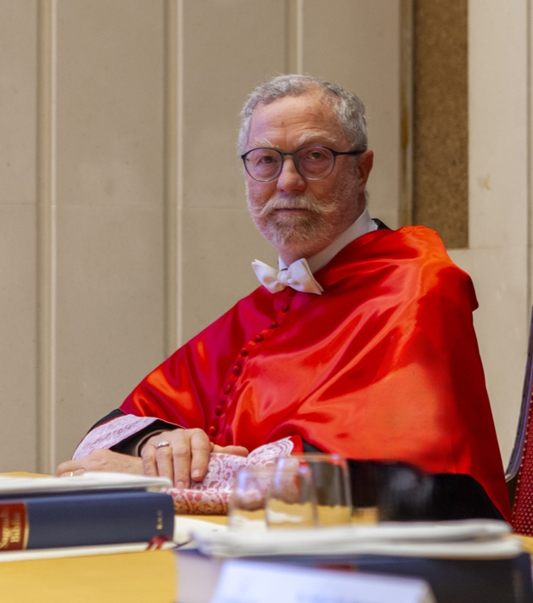 Un hombre mayor con barba y gafas vestido con una toga roja y una camisa blanca con corbata de lazo se sienta en un espacio interior posiblemente durante una ceremonia formal.