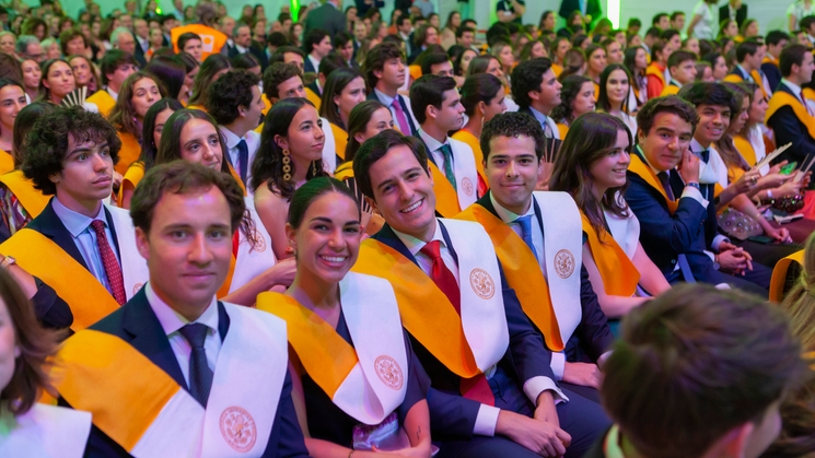 A group of young graduates wearing academic sashes and robes at a graduation ceremony, smiling and looking towards the stage.