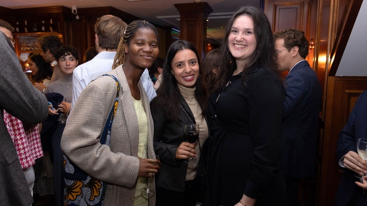 Three women smiling at the camera in a crowded event with drinks in their hands.