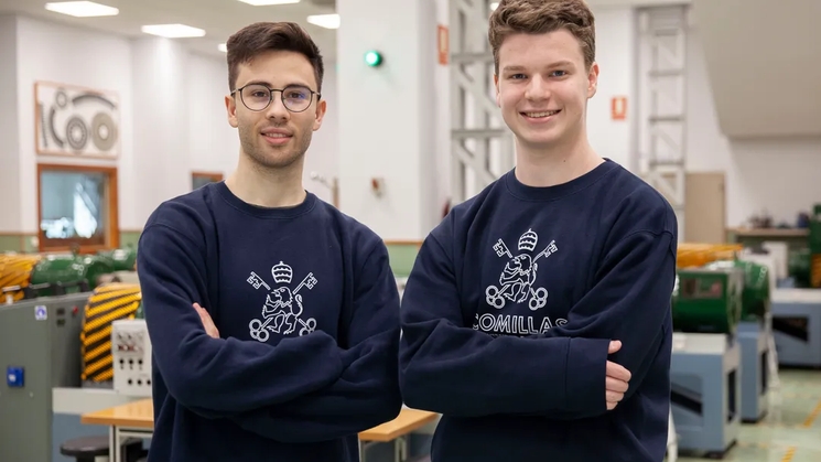 Dos jóvenes posan sonrientes en un taller con maquinaria al fondo.
