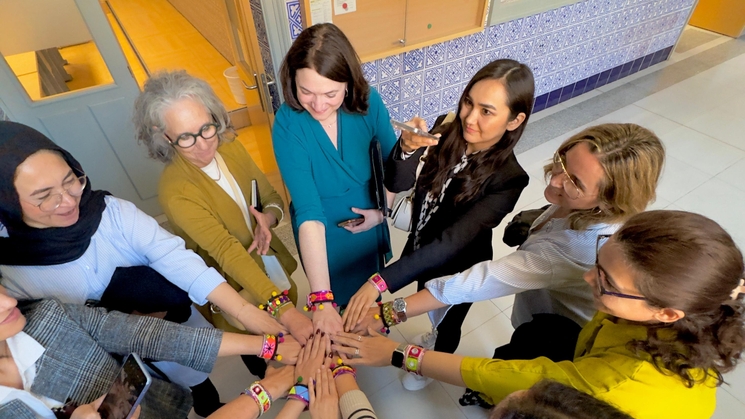 A group of women in professional attire joining hands in a circle from above, symbolizing teamwork and unity.