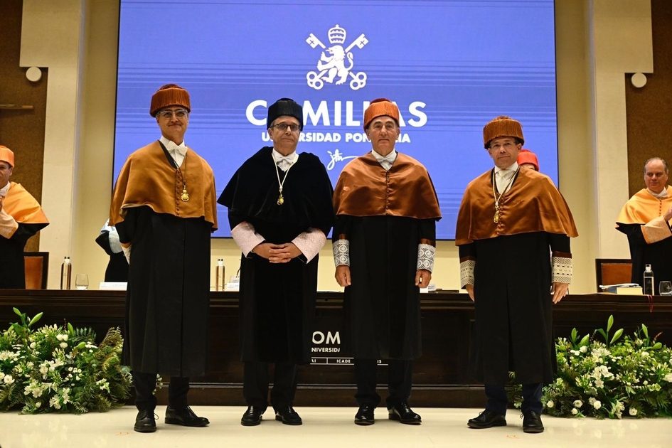 Four individuals in traditional academic attire standing in front of a banner with the Comillas University logo at a formal event.