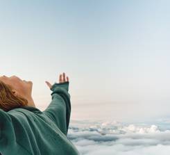 A person in a green hoodie standing with arms outstretched above the clouds, expressing freedom and joy.
