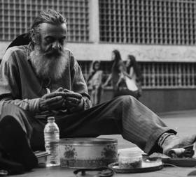 An elderly bearded man sitting on the ground, looking at something in his hands with his belongings scattered around him.