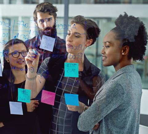 A group of four diverse colleagues brainstorming with sticky notes on a glass wall in a modern office.