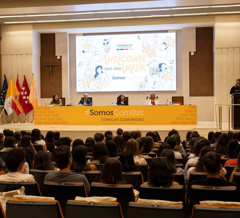 People attending a welcome event in an auditorium with speakers seated at a panel table under a banner.