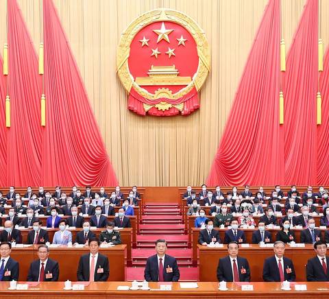 La imagen muestra una asamblea formal con una gran cantidad de personas sentadas en un auditorio decorado con cortinas rojas y el emblema de China en la parte superior.