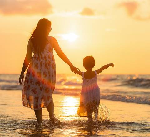 Una madre y su hija caminan de la mano por la orilla del mar al atardecer.