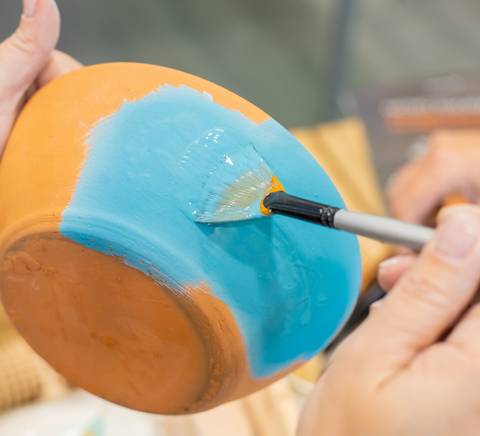 Una persona está pintando de azul una vasija de cerámica que ya tiene una base naranja.