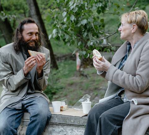 Dos personas, una con pelo largo y barba y otra con corte pixie, comiendo sándwiches en un parque, sentadas en un banco de piedra.