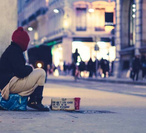 A person sitting on the sidewalk at night, with a cup presumably for collecting money and pedestrians in the background.