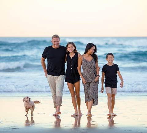 Una familia camina por la orilla de la playa al atardecer acompañada de un perro.