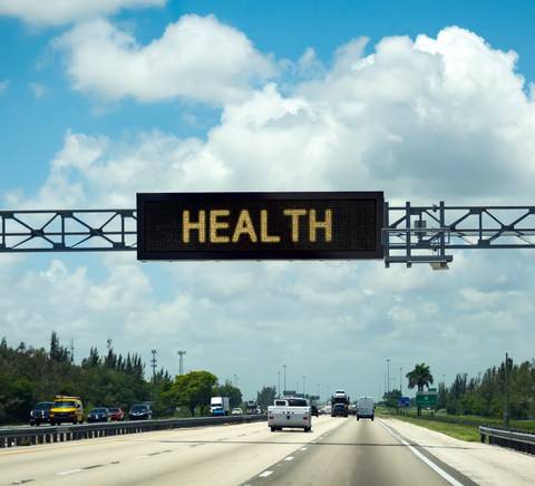 An electronic road sign over a highway displaying the word 'HEALTH' under a blue sky with clouds.