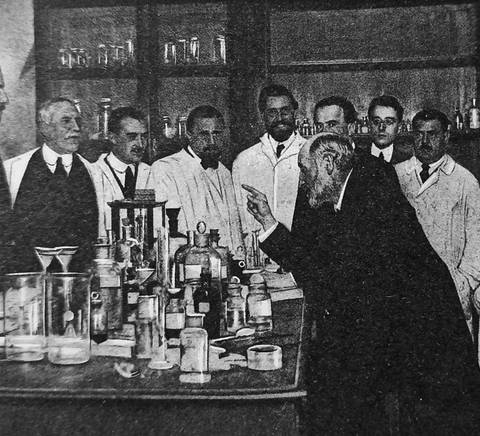 Un grupo de hombres observa una demostración en un laboratorio antiguo lleno de frascos y equipamiento científico.