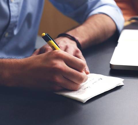 Una persona está escribiendo notas en un papel mientras usa una computadora portátil.