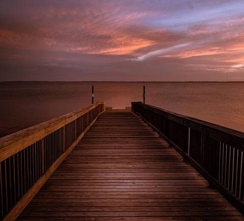 Un muelle de madera se extiende sobre un lago al atardecer, rodeado de cielos nublados y tonos rojizos.