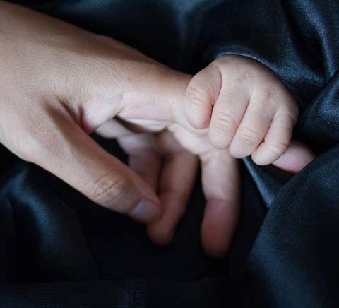 Una mano adulta sostiene la pequeña mano de un bebé sobre una tela oscura.