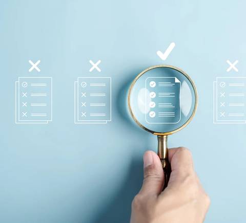 Una mano sostiene una lupa sobre documentos que muestran marcaciones y un documento con una marca de verificación.