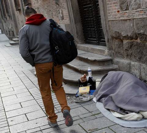 Una persona camina por la calle mientras otra está acostada con una manta en el suelo.