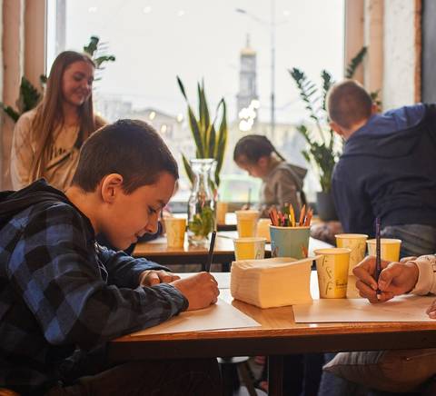 Un grupo de niños está dibujando en una mesa dentro de un café, rodeados de coloridos materiales de arte.