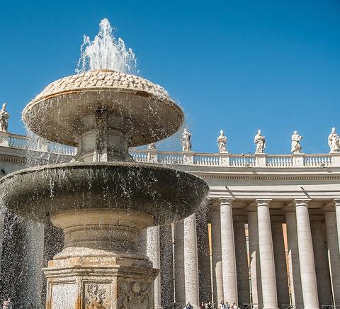 Una fuente decorativa en una plaza con columnas y estatuas al fondo bajo un cielo azul.