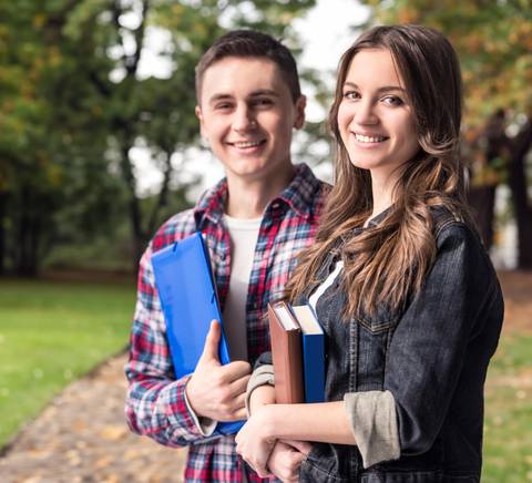 Two young students smiling and holding books in a park.