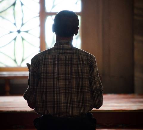 Un hombre está de espaldas, en posición de oración, dentro de un lugar iluminado por la luz que entra por una ventana decorativa.