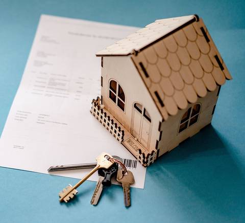 Una maqueta de casa de madera junto a un conjunto de llaves y un documento sobre fondo azul.