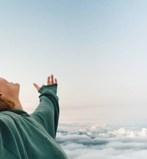 A person in a green hoodie standing with arms outstretched above the clouds, expressing freedom and joy.