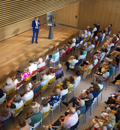 A man in a blue suit is giving a presentation to an audience seated in colorful chairs in a modern hall.