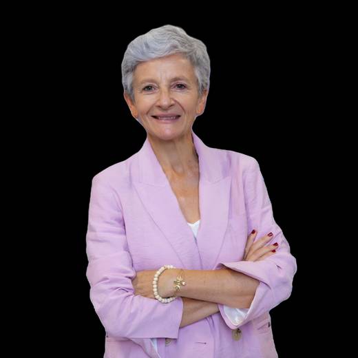 A senior woman in a pink blazer stands with arms crossed, smiling against a black background.