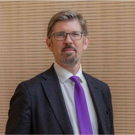 A portrait of a middle-aged man with glasses wearing a striped suit and a purple tie, standing in front of a wooden slatted wall.