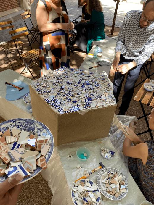 Un grupo de personas trabajando en la creación de un objeto decorativo con fragmentos de cerámica en una mesa al aire libre.