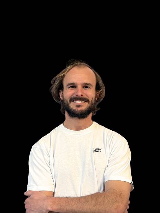 Un hombre sonriente con camiseta blanca de la marca Vans posa con los brazos cruzados frente a un fondo negro.