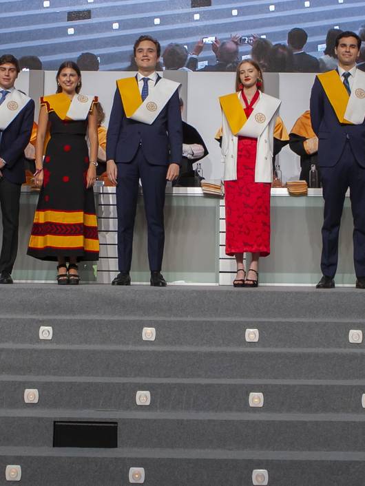 A group of eight young adults in graduation attire, standing on a platform.