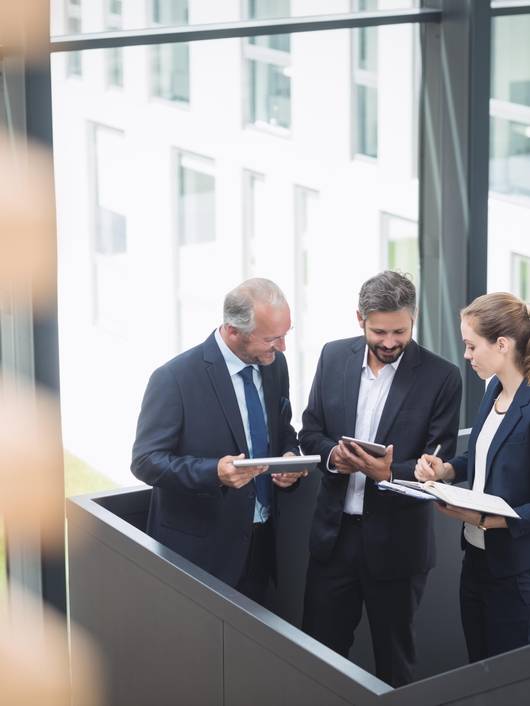 Three business professionals, two men and one woman, are discussing over documents in a modern office building.