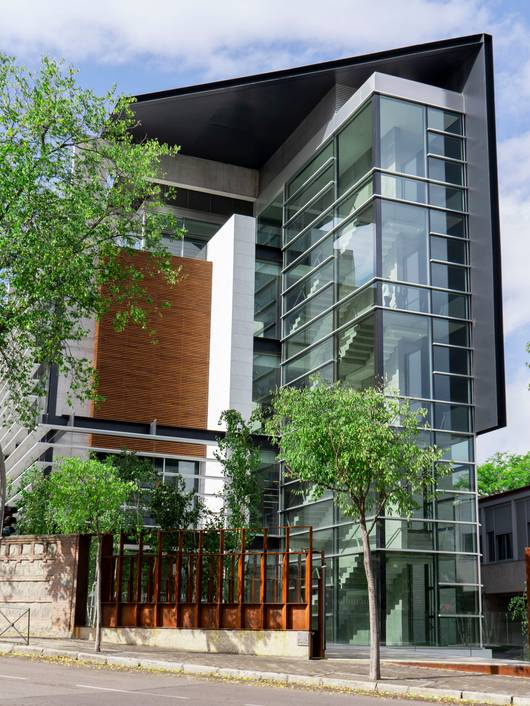 Modern glass building with orange accents on a city street lined with green trees.