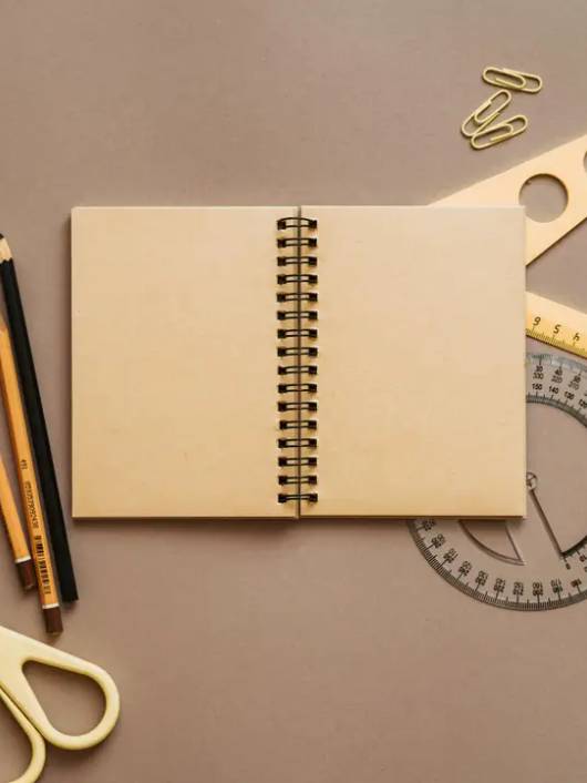 Una mesa de trabajo con un cuaderno vacío, lápices de colores, tijeras, un transportador y reglas.