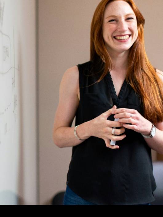 A joyful red-haired woman smiling and standing in a room with a whiteboard in the background.
