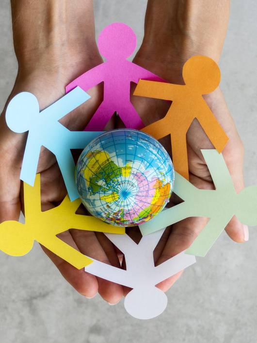 Unas manos sostienen un globo terráqueo rodeado de figuras de papel de colores que representan personas unidas.