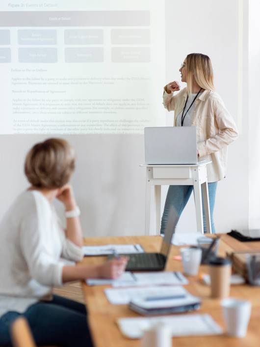 A woman standing and presenting at a workshop while another woman sits listening.