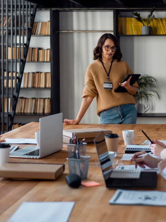 A young woman stands presenting in a boardroom meeting to two older colleagues who are taking notes.